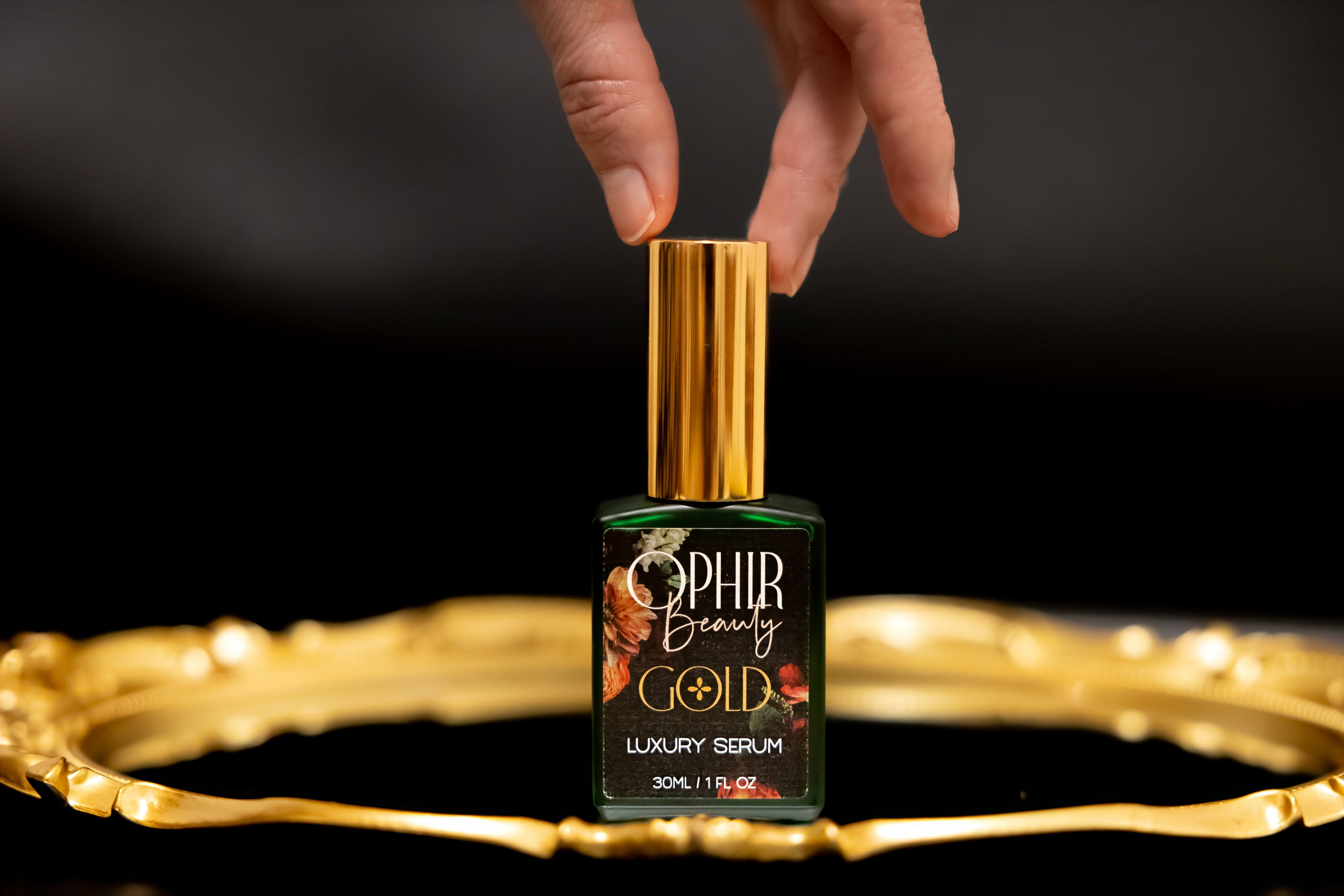 A woman's hand reaches for a green glass bottle of Ophir facial serum sitting on an ornate gilded tray.